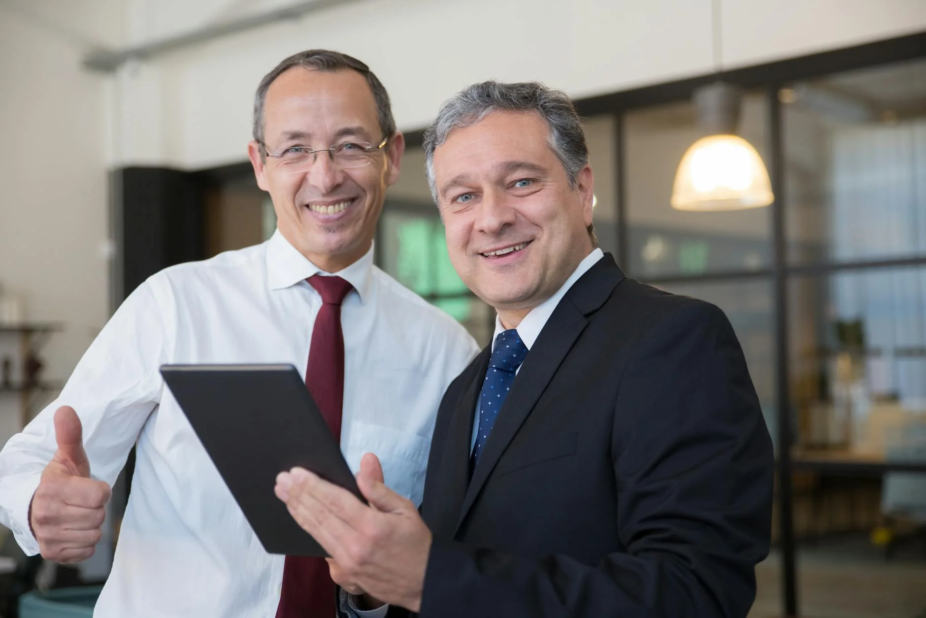 Two smiling businessmen in corporate attire discussing work on a tablet in a modern office.